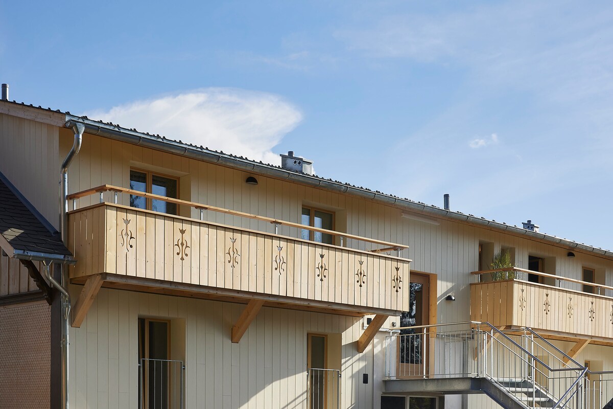 The exterior of the Gantrisch Lodge features a two-story wooden structure with balconies and decorative wood carvings. A metal staircase leads to the upper level, where large windows allow natural light to brighten the space. The clear blue sky serves as a backdrop.