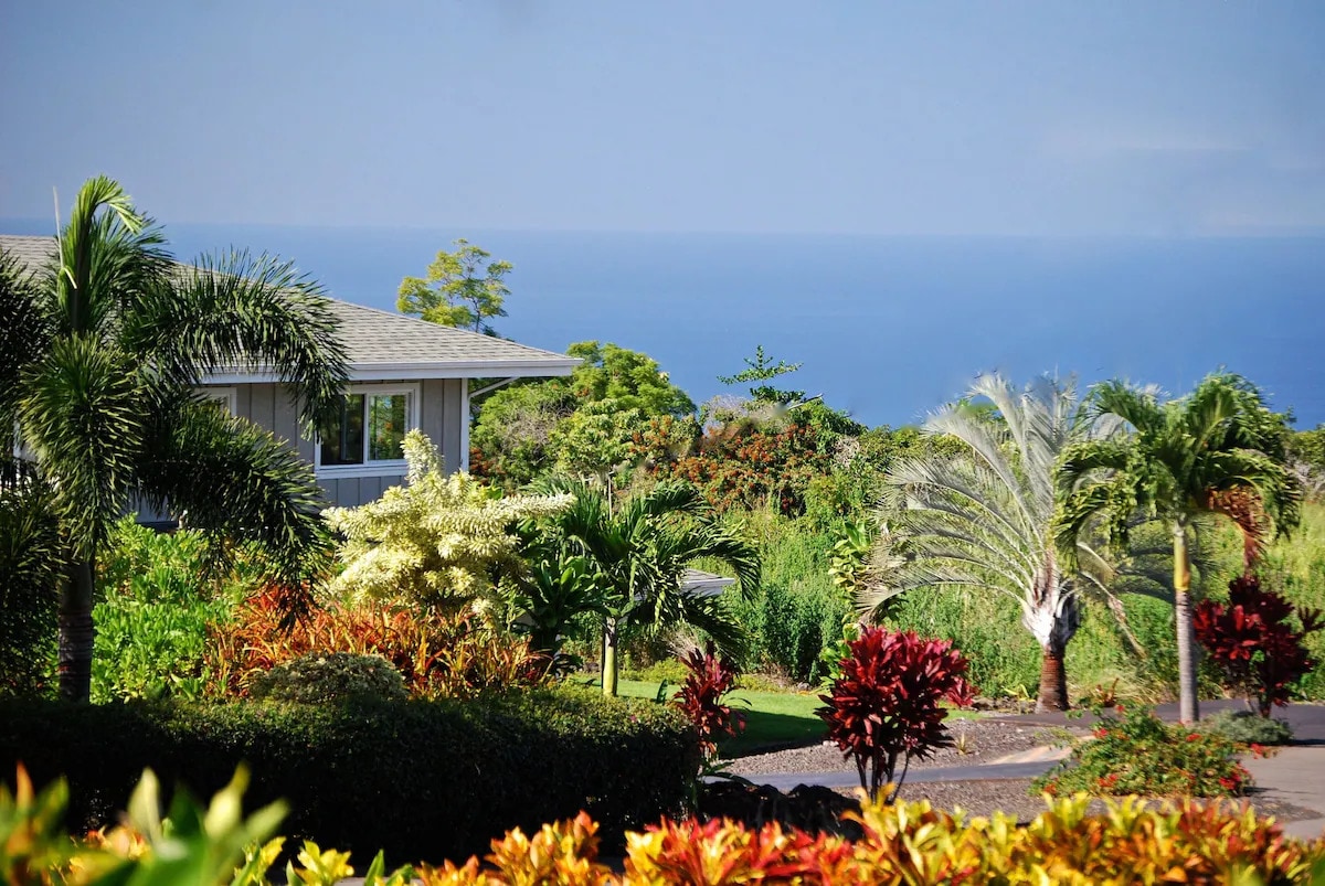 Lush tropical foliage surrounds a well-maintained property, with vibrant plants in shades of green and red. In the background, the ocean is visible, complemented by a clear blue sky. The serene landscape creates a tranquil setting for relaxation and exploration.