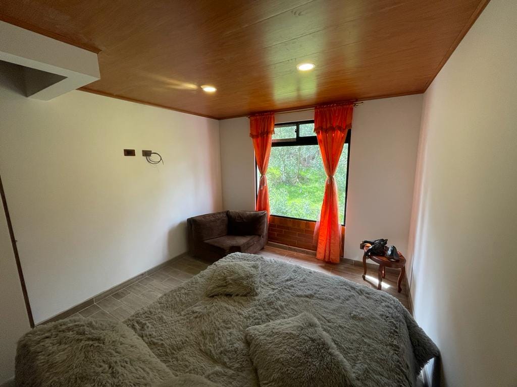 A cozy bedroom features a large bed covered in a soft, textured blanket. Natural light filters through a window dressed with orange curtains, illuminating an inviting corner with a dark armchair and a small table.