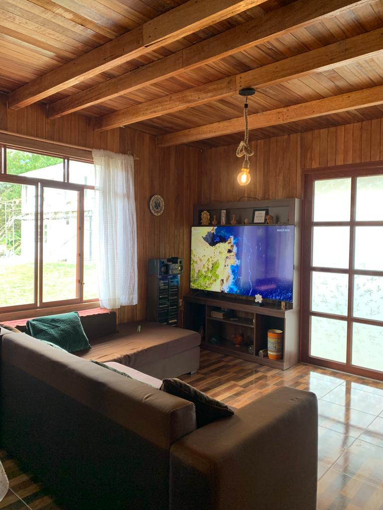 A cozy living room features a brown sectional sofa and a large television mounted on the wooden wall. Natural light enters through a window adorned with sheer white curtains, illuminating the rich wooden beams of the ceiling and the warm wooden floor.