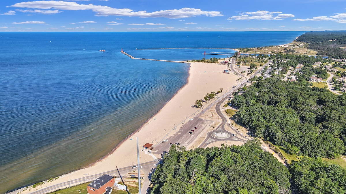 An expansive view of a sandy beach stretches along the coastline, meeting a calm blue sea. A jetty extends into the water, and the shoreline is bordered by lush greenery and pathways, offering a serene and inviting atmosphere for outdoor activities.