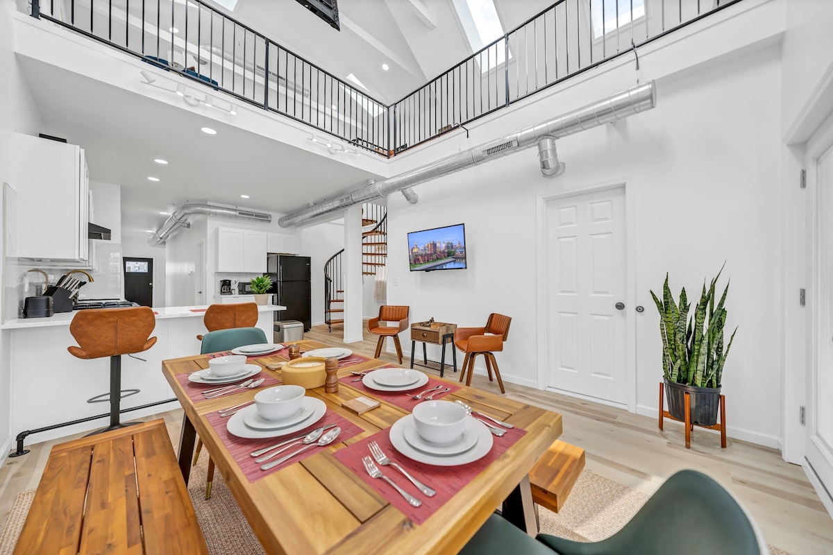 The dining area features a large wooden table set with dishes and silverware, surrounded by wooden and black chairs. A spiral staircase is visible in the background, leading to an upper level. Modern appliances and a large window are seen in the kitchen space.