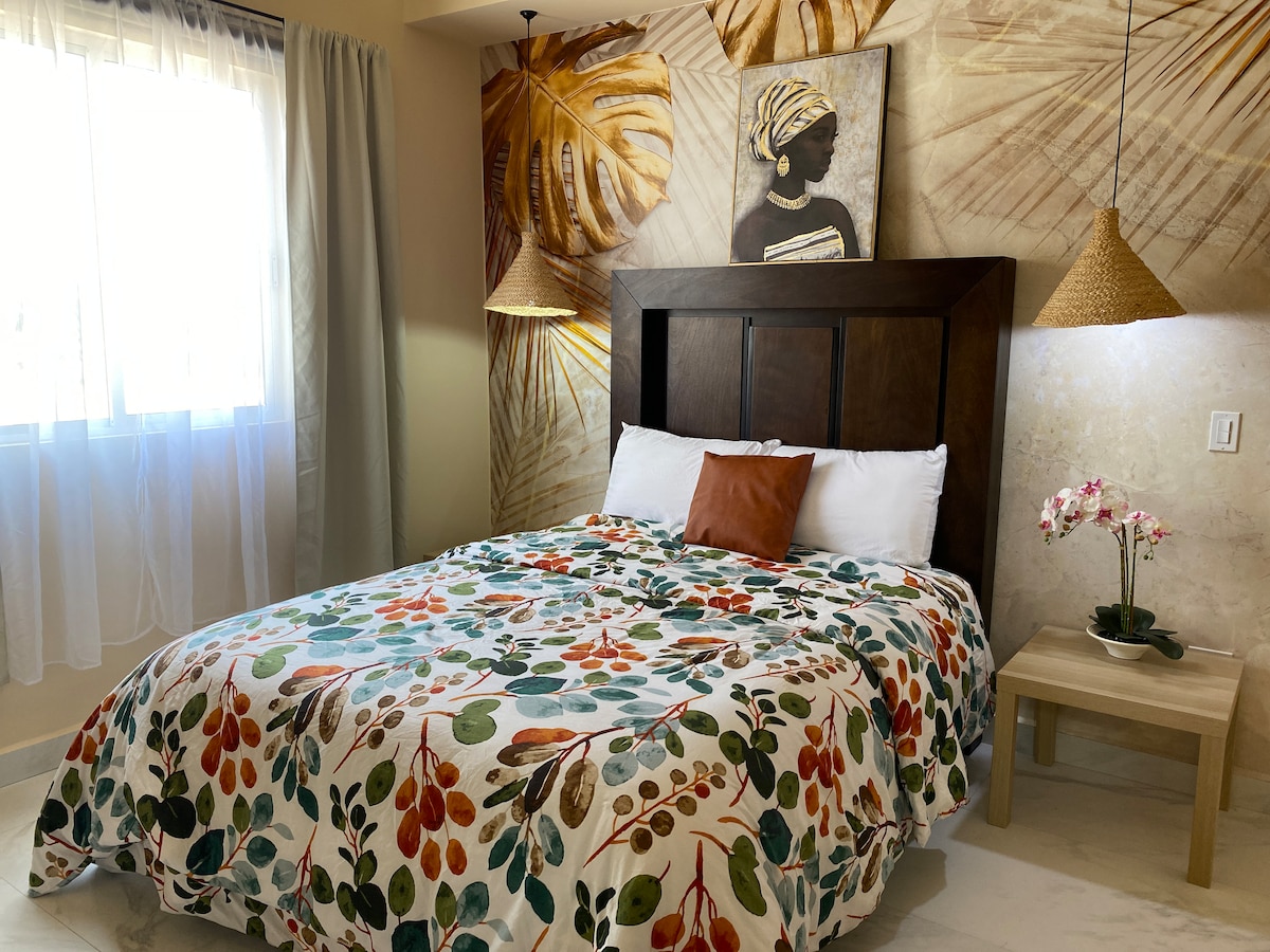 A cozy bedroom features a spacious bed with a floral patterned blanket, complemented by a decorative pillow. Natural light filters through sheer curtains, illuminating the warm-toned wall adorned with tropical motifs and artwork. Two woven pendant lights hang elegantly above the bed.