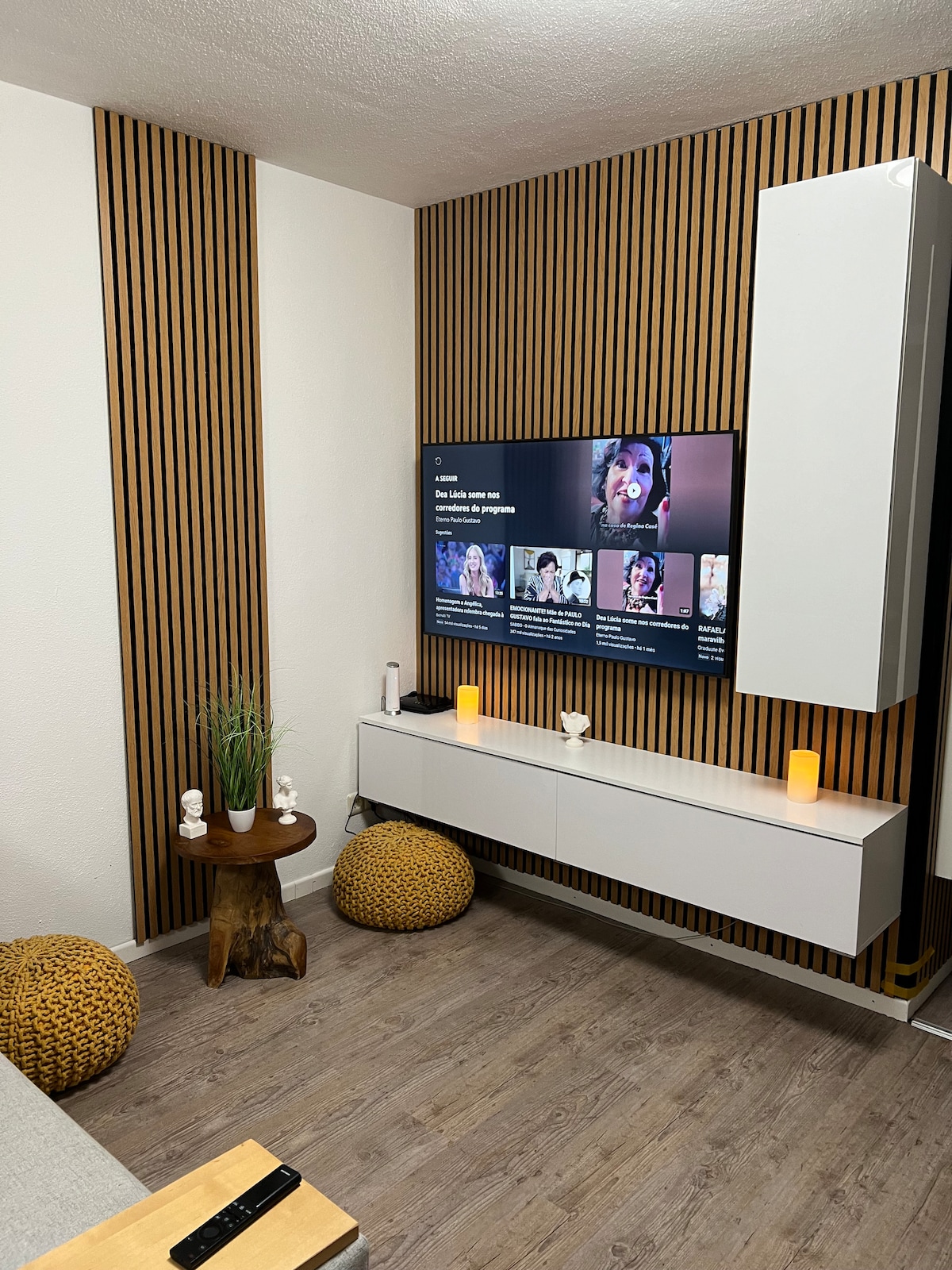 A modern living area is presented with wooden accent walls and a sleek media console. A large television is mounted on the wall, while two textured poufs provide additional seating. Soft lighting from strategic candles enhances the cozy ambiance.