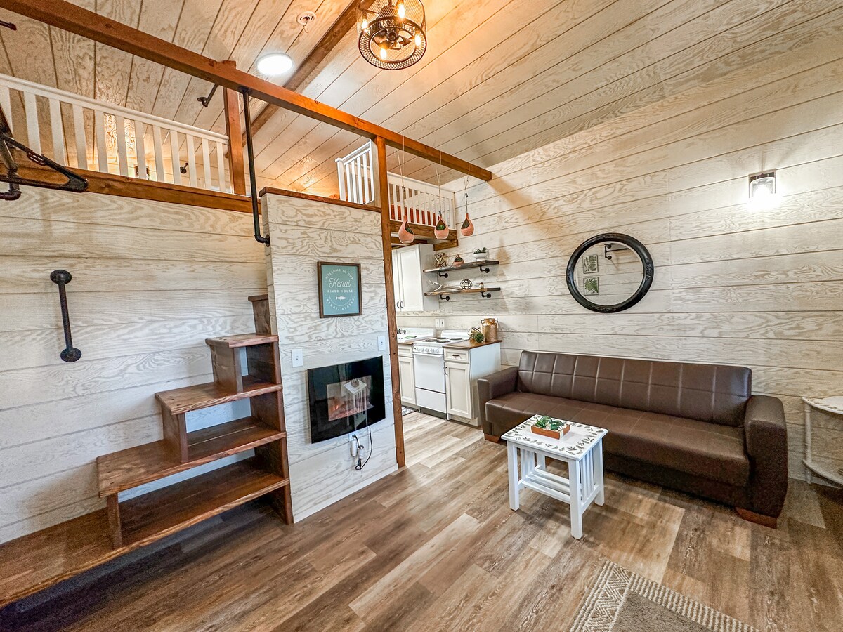 The interior features a cozy living space with a comfortable brown couch and a small white coffee table. Stairs lead to a sleeping loft above, accentuated by warm wooden beams. A fully equipped kitchen is visible in the background, alongside a decorative wall mirror.