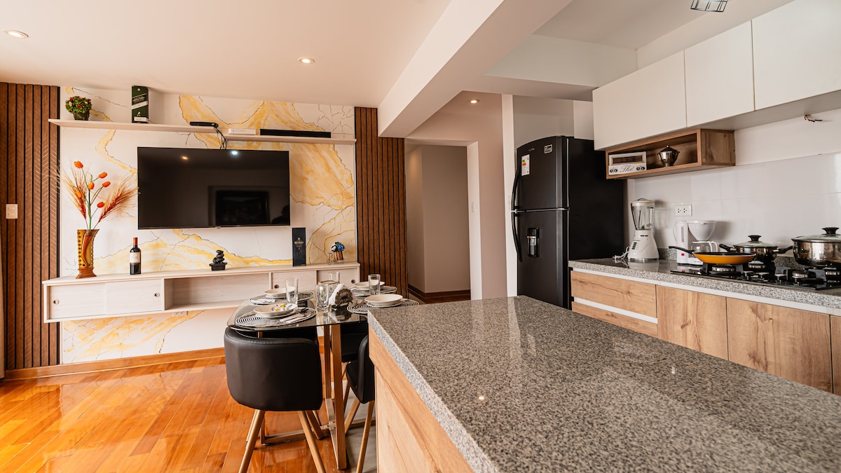 A modern kitchen and living area are showcased, featuring sleek cabinetry and granite countertops. A dining area for four is visible, along with a large television mounted on the wall. Appliances such as a refrigerator and blender are neatly arranged, complementing the open layout.