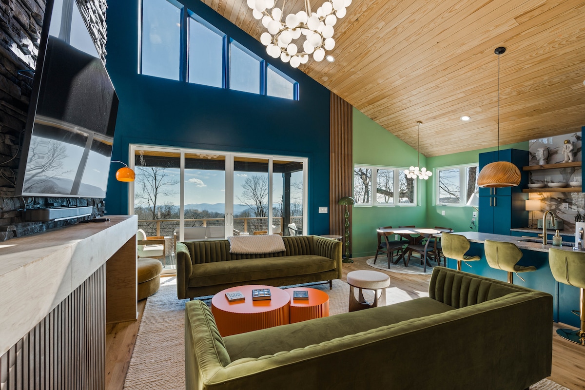 A spacious living area is defined by high vaulted ceilings and large windows, allowing natural light to fill the room. Green velvet sofas are positioned around a circular coffee table, while stylish seating at the kitchen counter enhances the inviting space.