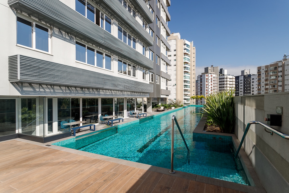 A modern outdoor swimming pool is seen alongside a sleek building. Lounge chairs are positioned along the pool's edge under bright sunlight, and plants provide a touch of greenery. The clear water reflects the blue sky, enhancing the inviting atmosphere of the space.
