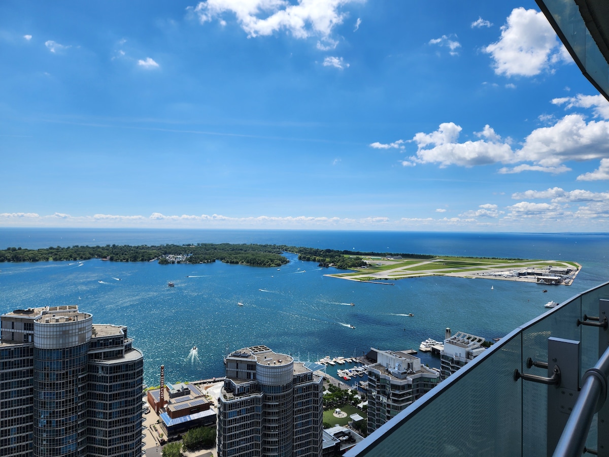 A scenic view showcases Lake Ontario's expansive waters, dotted with boats and framed by lush greenery. The skyline features modern buildings and a clear blue sky with soft clouds, offering a serene contrast against the vibrant lake.