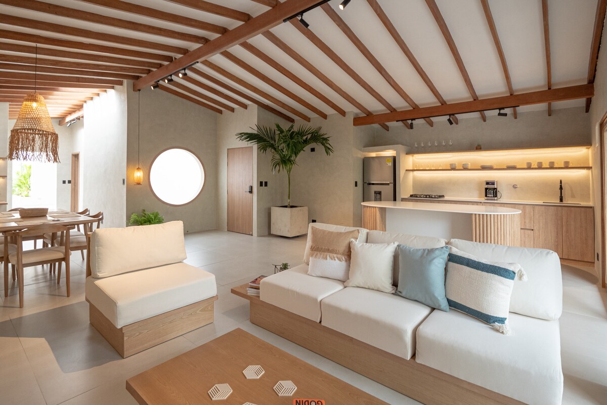 An open living space is presented with a light-colored sofa adorned with various cushions. Natural wood accents and beams are featured in the ceiling. A circular window allows light to enter, while a dining area and kitchen are visible in the background.
