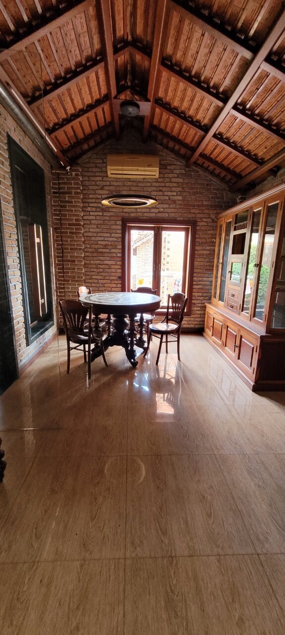 A dining area features a round wooden table surrounded by four chairs, set against a backdrop of exposed brick walls. High ceilings showcase wooden beams, and natural light streams through a window, illuminating the polished flooring.