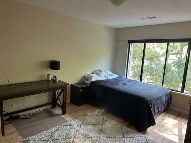 A quiet bedroom is displayed with a spacious bed positioned next to a nightstand. A desk with a lamp is situated nearby. Large windows invite natural light, offering views of tree foliage outside. The floor is covered with a light-colored area rug.