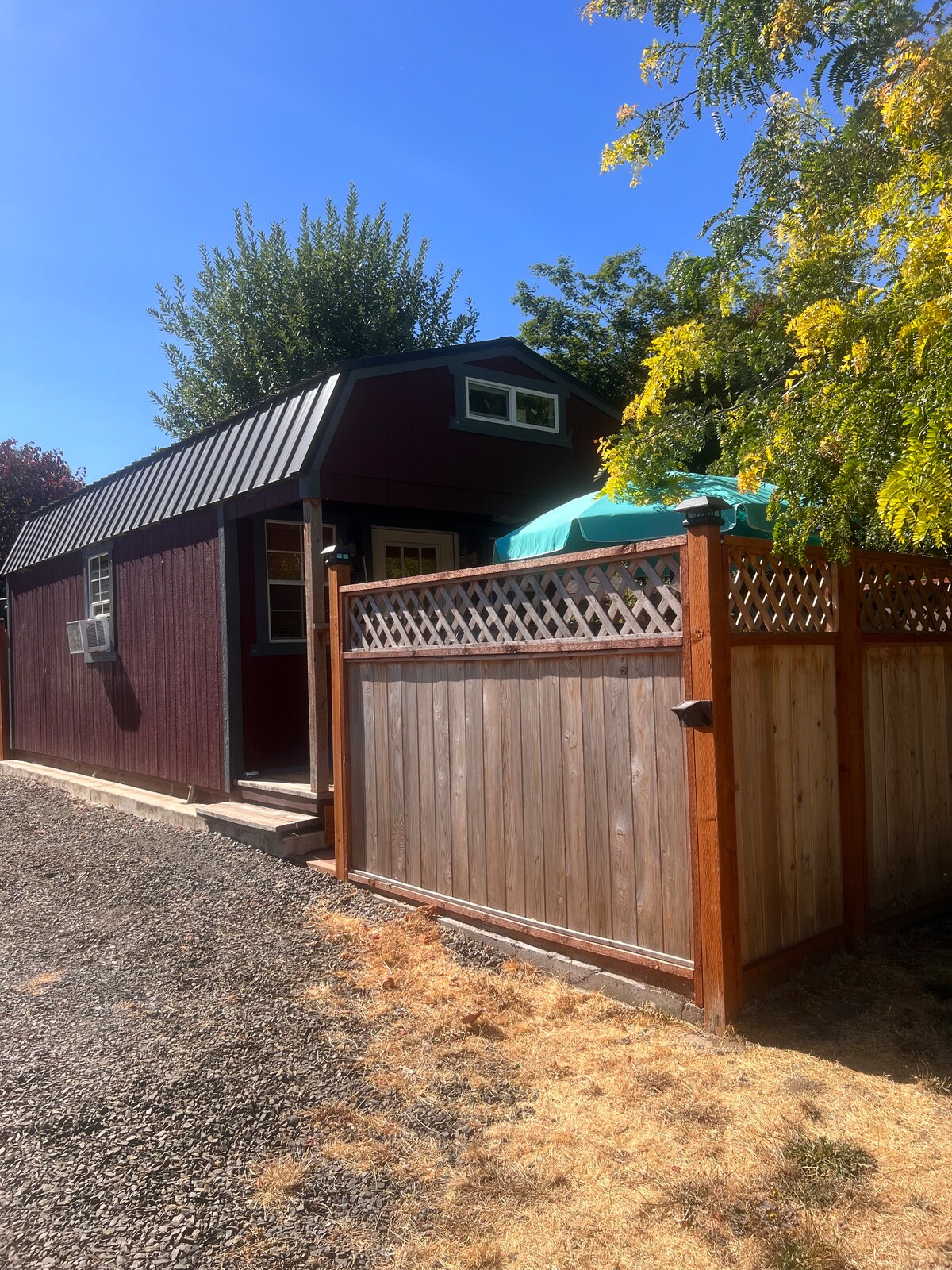 A cozy red cottage with a metal roof is surrounded by a wooden fence. Lush greenery frames the structure, and sunlight casts a warm glow on the gravel path leading to the entrance. A shaded area is visible, featuring a teal outdoor umbrella.