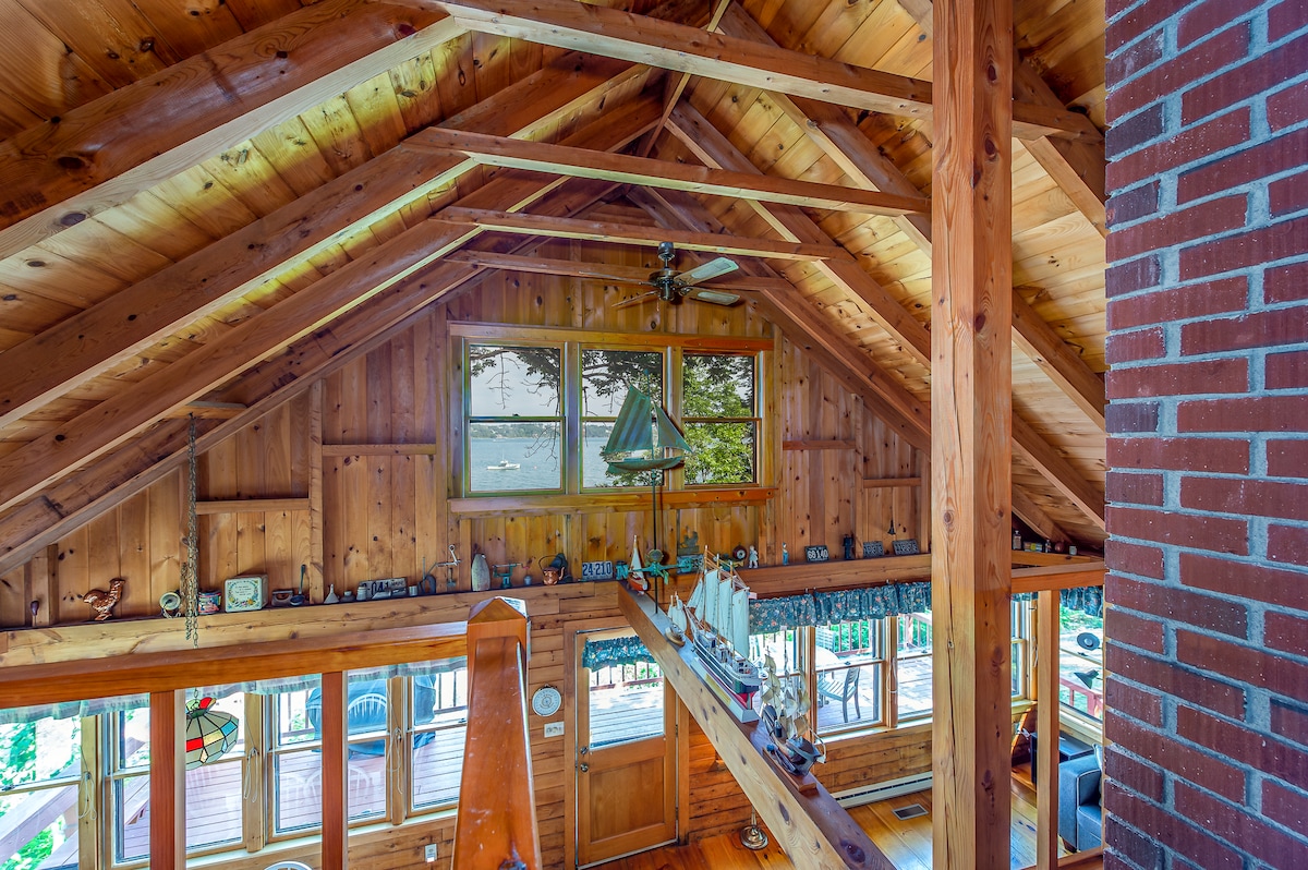 The interior features a high wood-paneled ceiling with exposed beams, lending an open feel. Large windows provide views of the waterfront, while natural light fills the space. Decorative items are placed along shelves and surfaces, contributing to the cabin's character.