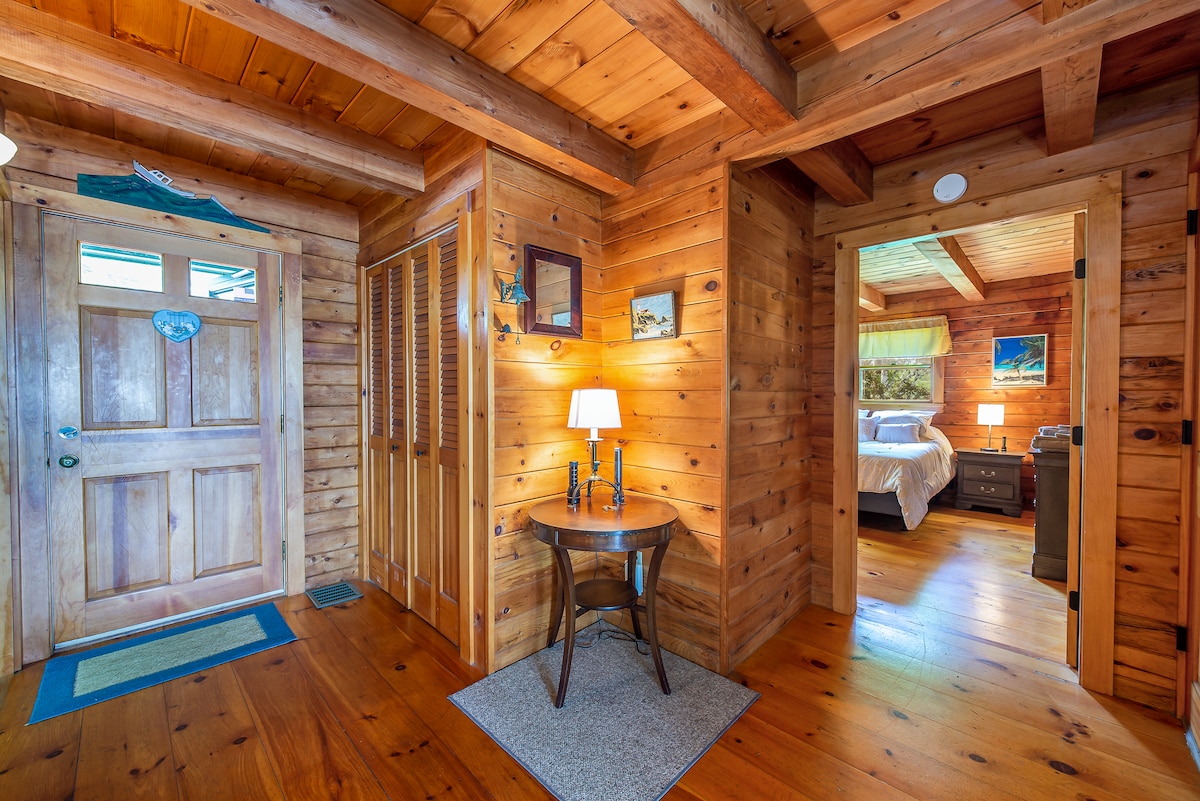 A welcoming entryway features wood paneling and warm lighting. A small table with a lamp sits beside the door, providing a cozy touch. Two arched doorways lead to separate rooms, enhancing the cabin's rustic charm, while a textured area rug adds comfort underfoot.
