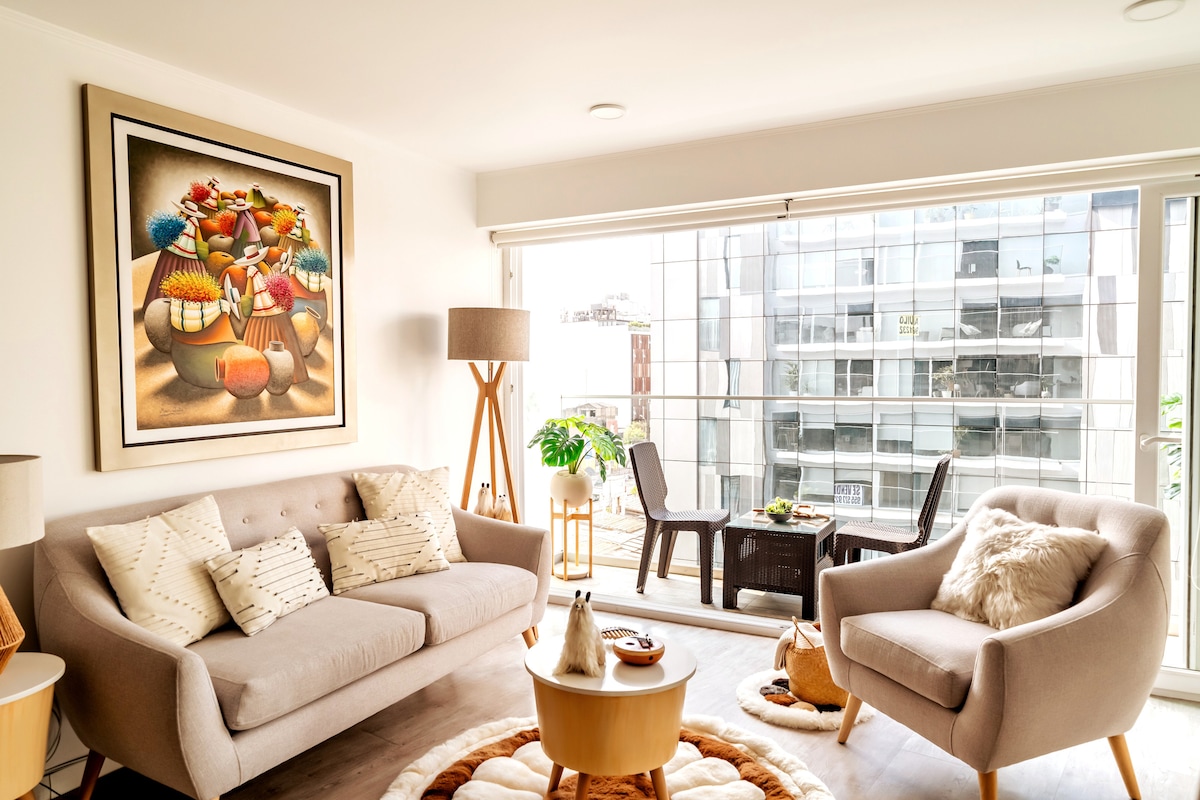 A cozy living space is featured, showcasing a light-colored sofa and armchair with cushions. A round wooden coffee table is positioned in front. The large windows provide natural light and views of the balcony, where outdoor seating is available. A vibrant artwork adorns the wall.
