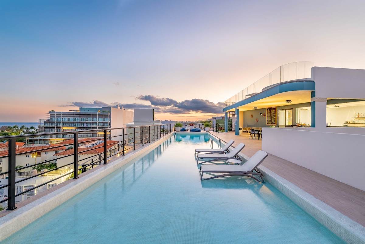 A rooftop pool stretches across the image, revealing a serene expanse of water reflecting the pastel colors of the sunset. Lounge chairs are positioned at the pool's edge, and an open terrace area features a casual dining space in the background, surrounded by a tranquil atmosphere.