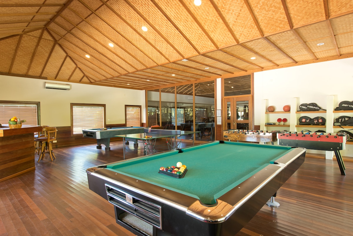 A spacious game room features a billiards table with a green felt surface and a variety of pool balls. In addition to the billiards area, foosball and table tennis tables are visible, along with a wooden dining table near large windows that invite natural light.