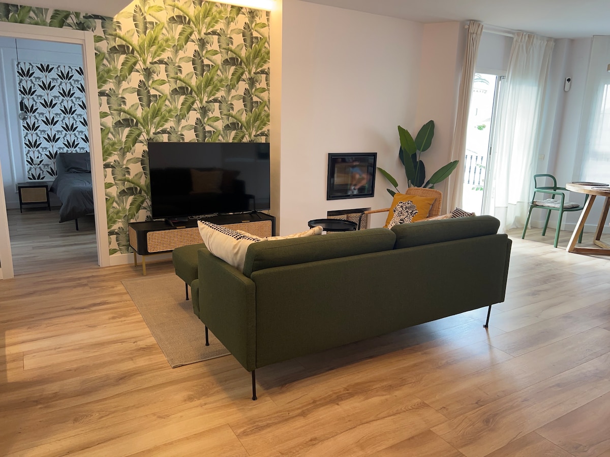 A modern living area is shown with a green sofa positioned in front of a television and fireplace. Bright light fills the space, featuring a palm leaf accent wall and wooden floors. A dining table with chairs is visible, enhancing the open layout.