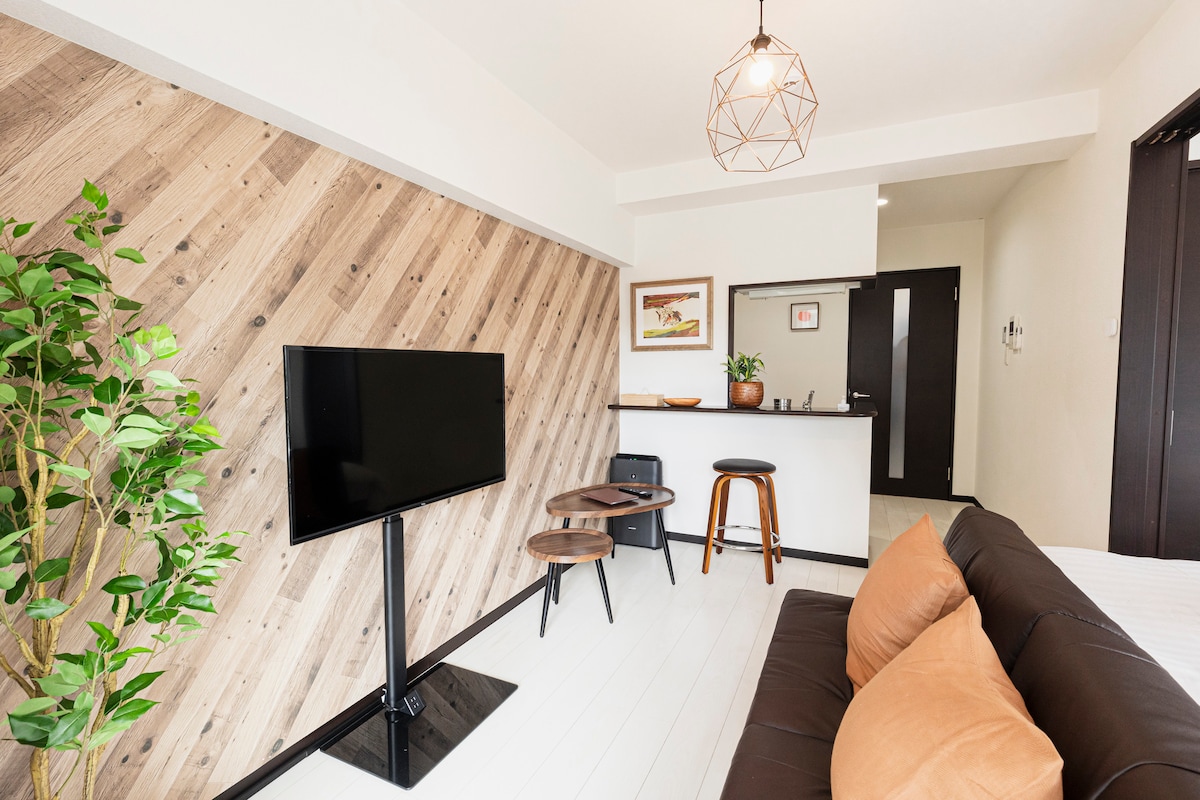 A modern living area is showcased with a smooth, light-colored floor and wooden accent wall. A flat-screen TV is mounted on a stand, facing a dark sofa with decorative cushions. Two small tables and a high chair are visible, with a kitchenette in the background.
