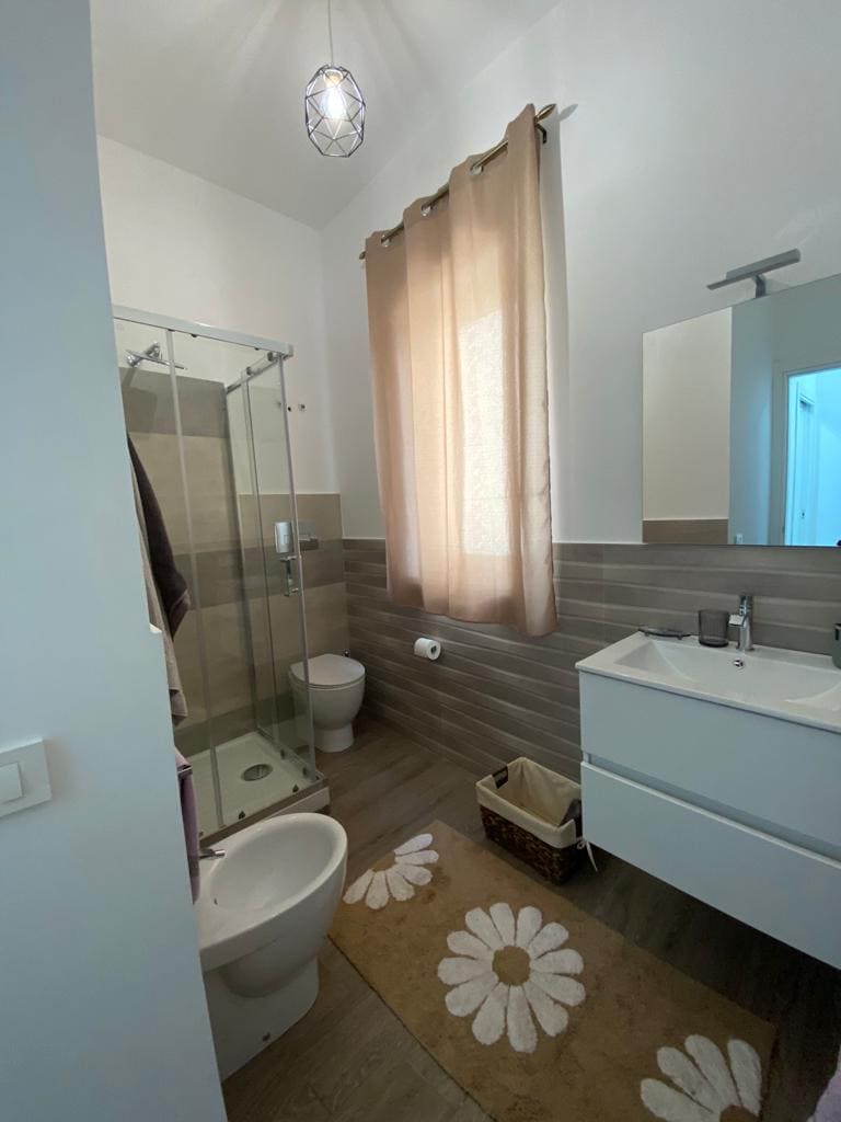 A modern bathroom features a glass shower with subtle tile accents, providing a spacious feel. Natural light filters through the sheer curtain, illuminating the light wood and tile flooring. A sleek vanity with a mirror and a towel basket are positioned against the wall.