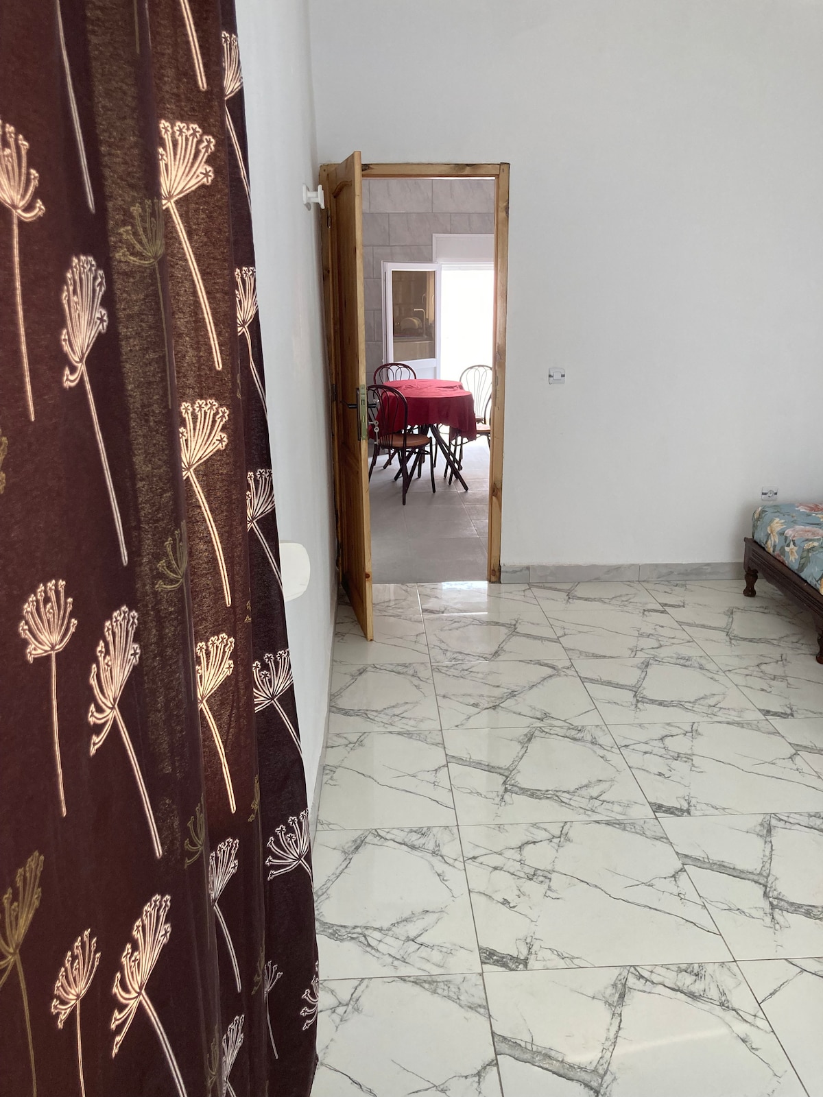 A well-lit hallway is highlighted by a patterned curtain and marble flooring. An open door leads to a dining area with a red tablecloth and several chairs. Natural light is visible from a nearby window, creating a bright and spacious atmosphere.