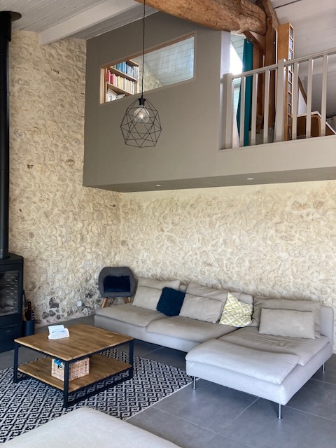 A spacious living area features a modern sectional sofa with neutral cushions, positioned near a wooden coffee table. Exposed stone walls provide a rustic backdrop, while a geometric pendant light hangs from the ceiling. A staircase leads to an upper level that includes a lofted area.