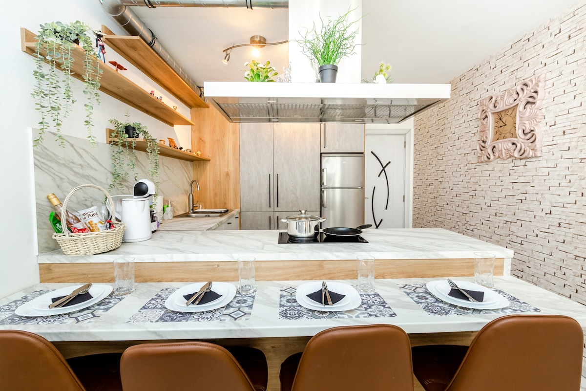 An open kitchen area is revealed, featuring a marble countertop and wooden shelving adorned with plants. A modern stove and stainless steel refrigerator are visible. A dining space with four brown chairs is set with plates, utensils, and glasses, creating an inviting dining environment.