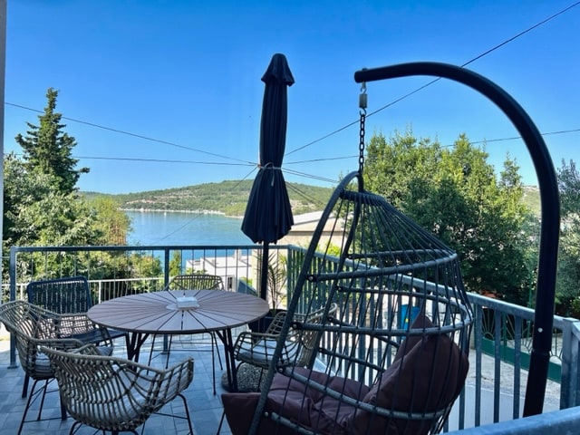 A spacious terrace is shown, featuring a round dining table surrounded by several wicker chairs. A hanging chair is positioned to one side, alongside a tall umbrella providing shade. Green trees and a scenic water view are visible in the background.