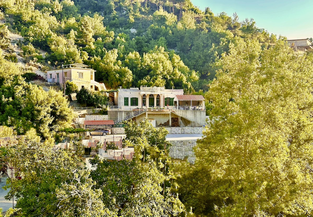 The property is nestled among lush greenery, featuring a light-colored, spacious structure with a prominent terrace. Surrounded by trees and hills, the scene reflects a serene countryside setting that invites relaxation and connection with nature.