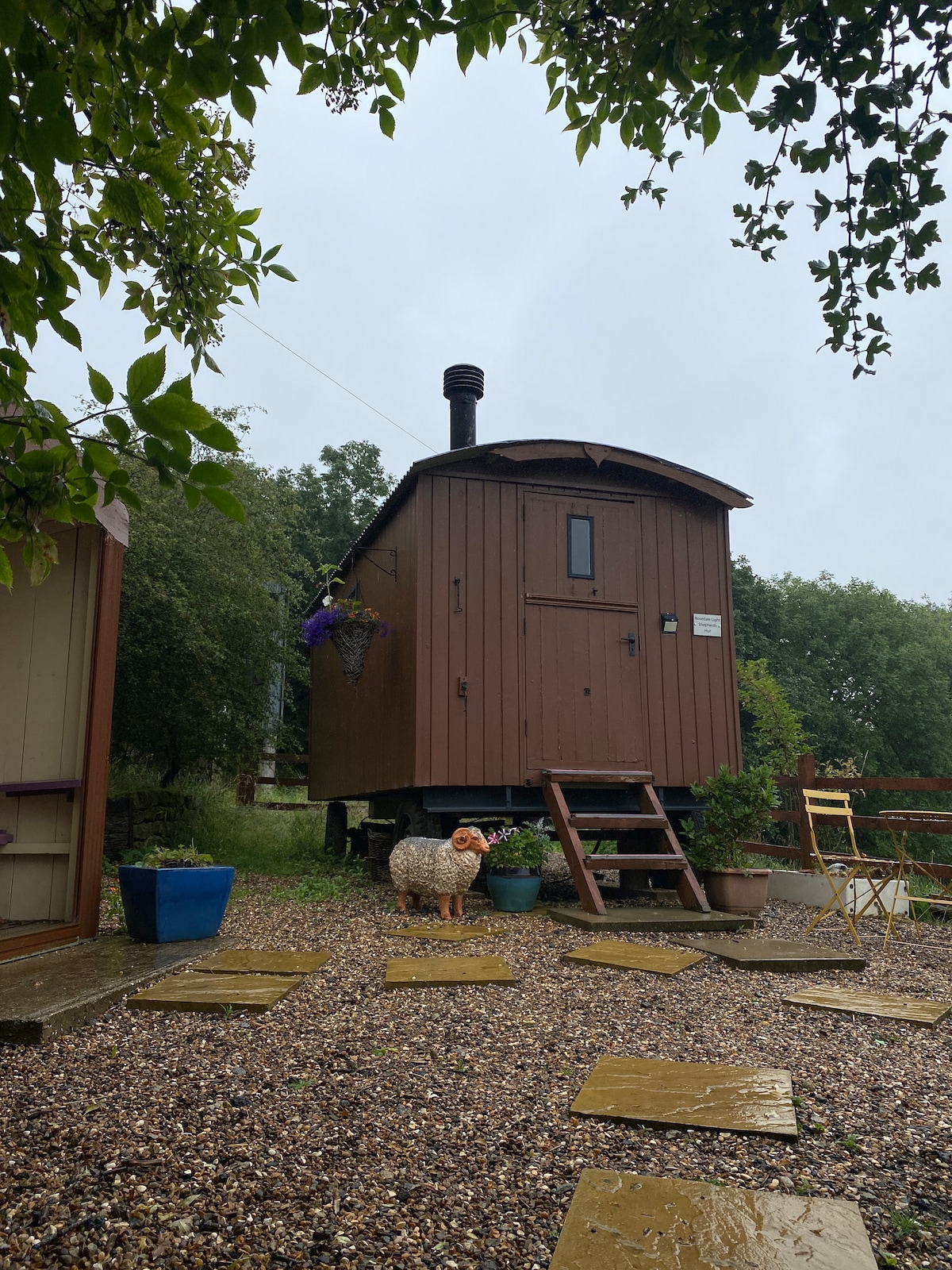 The exterior of a brown Shepherd's Hut is shown, nestled among greenery. A stone pathway leads to wooden steps, with a few decorative pots placed around. A sheep statue and colorful flowers are visible, enhancing the serene rural setting.
