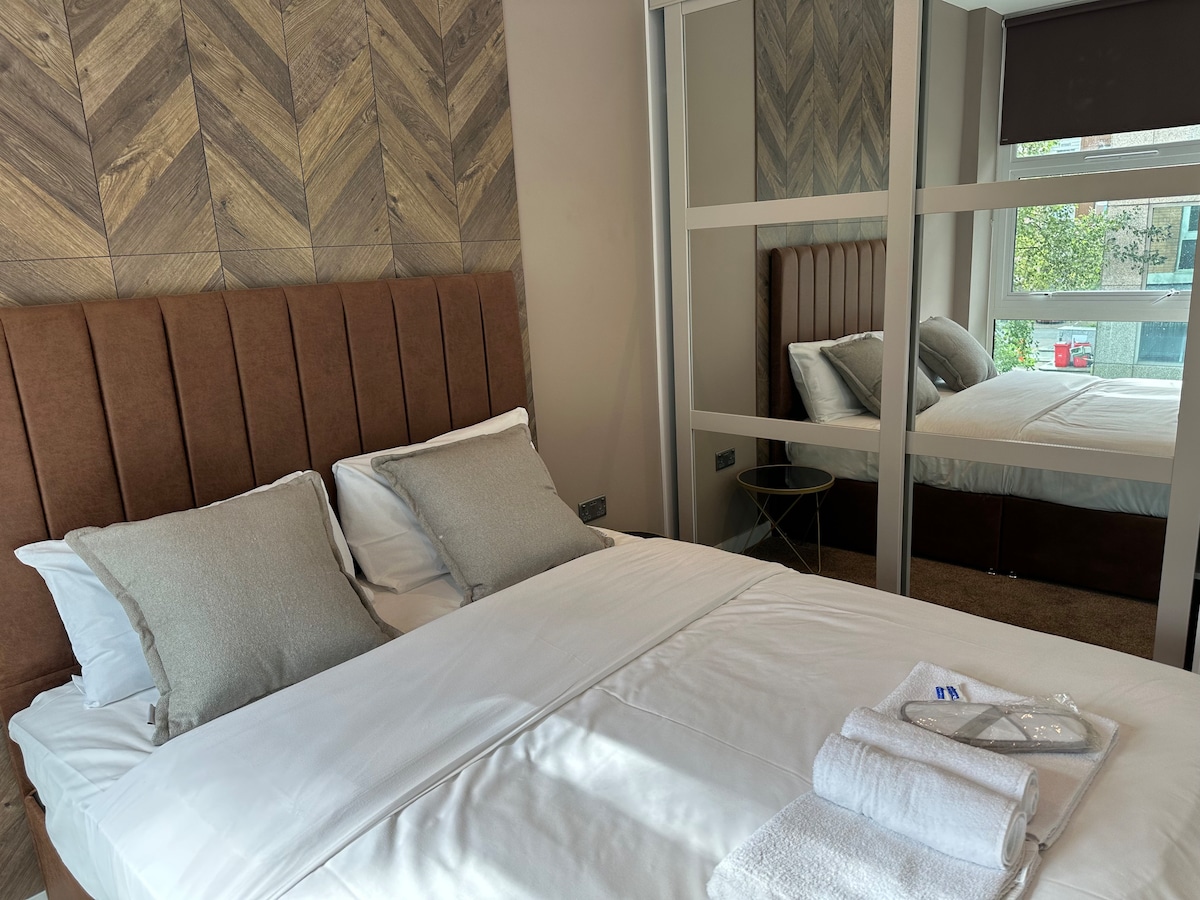 A well-appointed bedroom features a plush bed with dual pillows and crisp linens. A large mirror reflects the room's design, which includes a stylish wooden accent wall. A small side table and neatly rolled towels are also visible, enhancing the room's organized appearance.