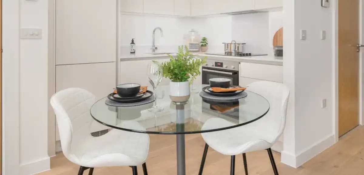 A modern dining area features a round glass table with a small potted plant at its center. Two white chairs are positioned around the table, which is set with black plates, silverware, and glasses, providing a clean and inviting space for meals.