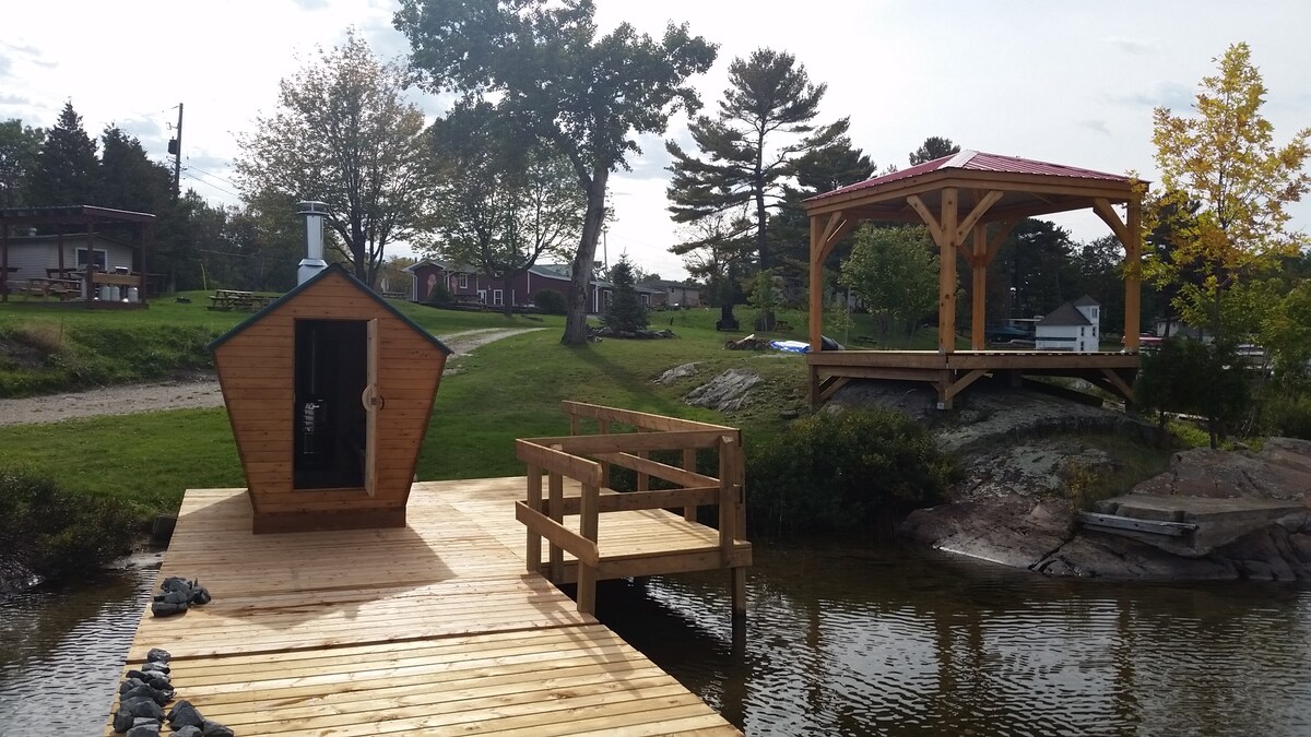A wooden dock extends over the water, featuring a small, rustic sauna structure. Nearby, a gazebo with a red roof offers shaded seating. Lush green grass surrounds the area, with trees in the background and additional cabins visible in the distance.
