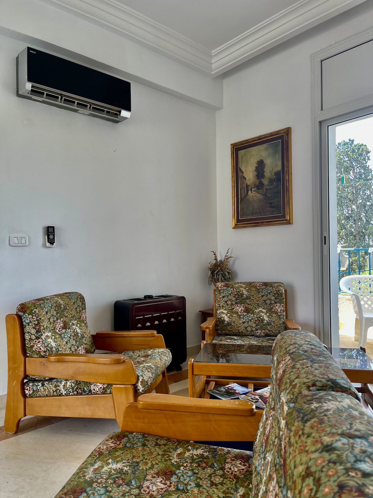 A welcoming living area is presented with wooden chairs and a small table, featuring floral upholstery. An air conditioning unit is mounted on the wall, while a painting adds a touch of decor. Light streams in through the partially open door leading to a balcony.