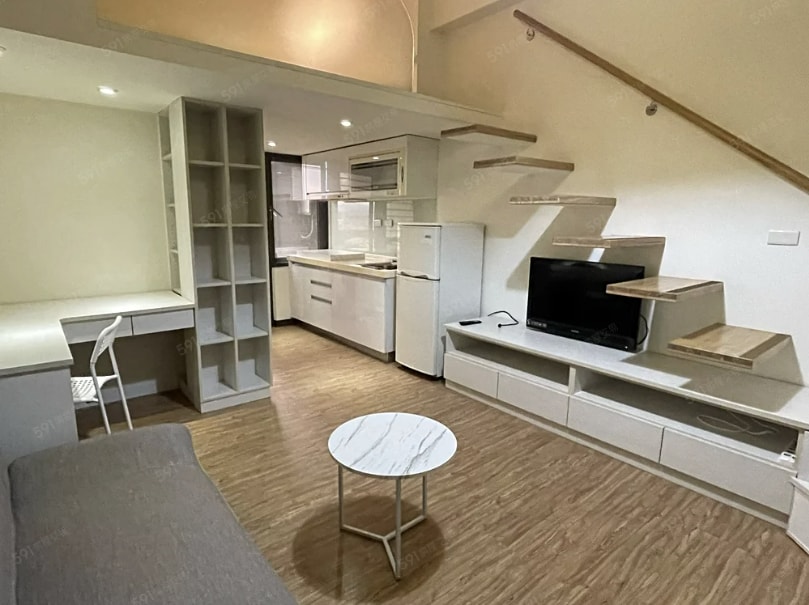 An open living area features a compact kitchenette, a refrigerator, and a television on a media console. A small white table is centered on wood flooring, and a staircase leads to an upper level. Natural light illuminates the space through a window.