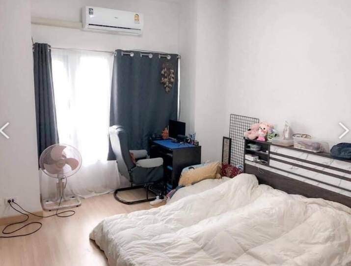 A well-lit bedroom features a spacious area with wooden flooring. Two bedding sets are arranged on the floor, complemented by a desk with a chair in the corner. A fan and a window with dark curtains are also visible, providing ventilation and natural light.