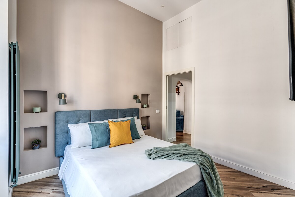 A spacious bedroom features a fitted bed with neutral bedding and decorative pillows in green and yellow. Soft lighting is provided by wall-mounted lamps. The room is complemented by wooden flooring and minimalistic shelving with decorative items, creating a calm and inviting environment.