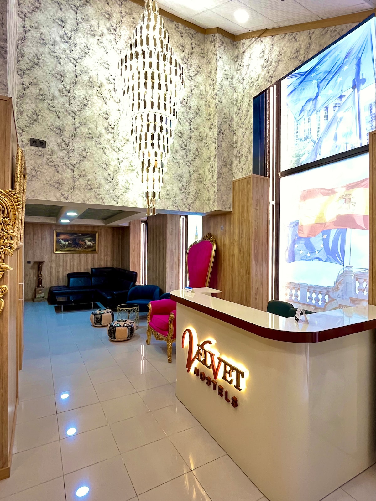 The reception area features a sleek front desk with the 'Velvet Hostels' logo prominently displayed. A modern chandelier hangs from a high ceiling, while large windows allow natural light to illuminate the space. Comfortable seating is provided, along with decorative elements enhancing the atmosphere.