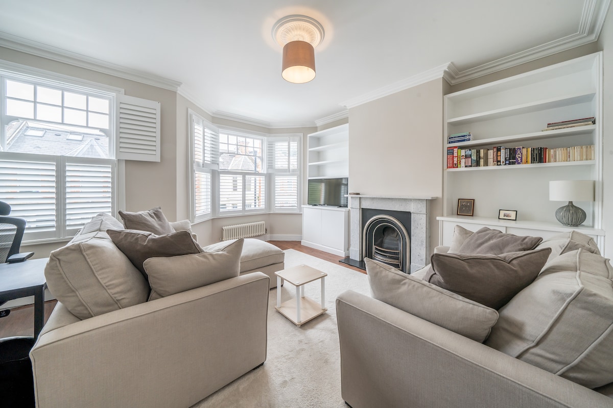 A spacious living area features soft beige sofas positioned around a small coffee table. Natural light enters through large bay windows, showcasing a light-colored rug. A fireplace is situated against the wall, while built-in shelves hold books and decorative items.