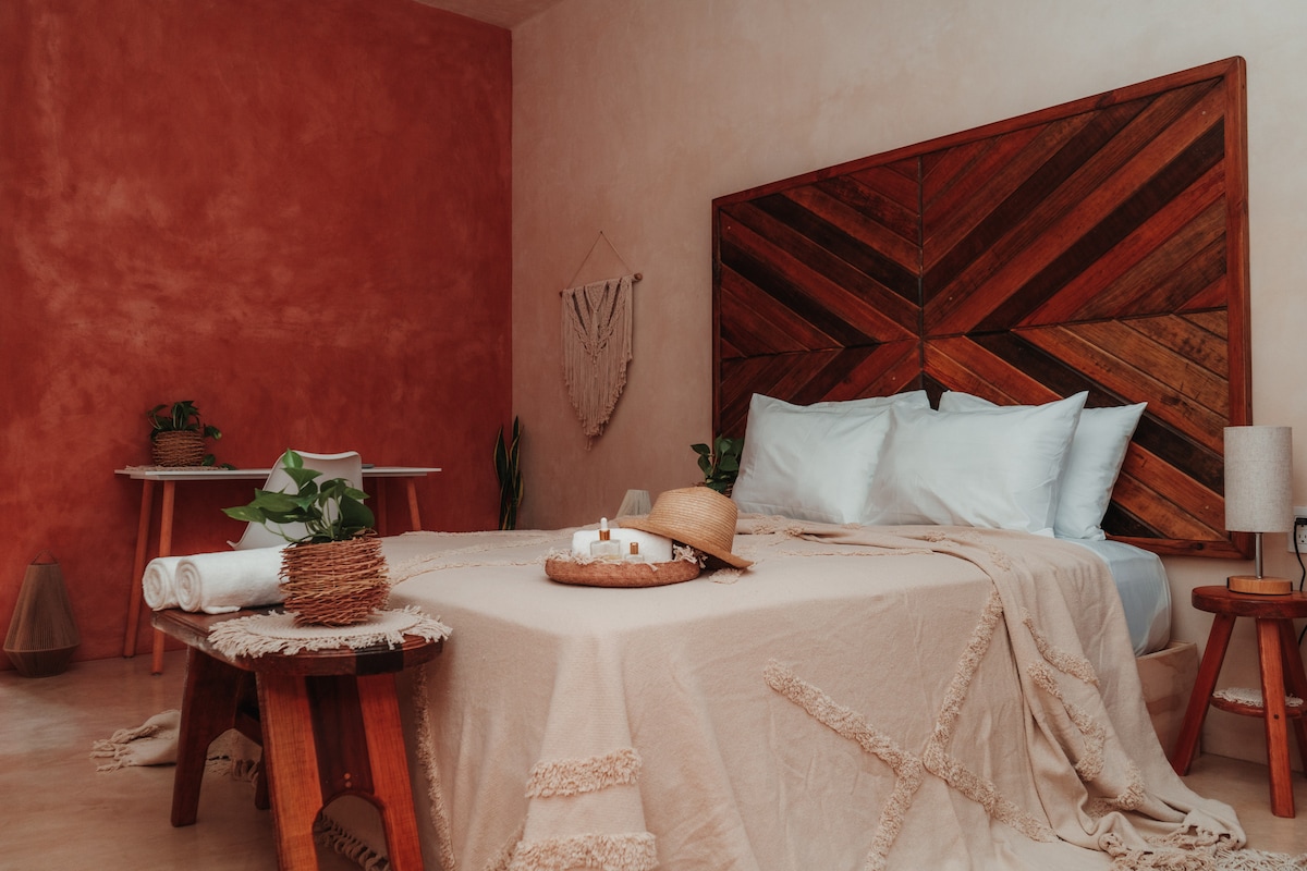 A serene bedroom features a spacious bed with a neutral blanket, flanked by wooden side tables. A warm-toned wall adds character, while indoor plants and decorative items enhance the calming ambiance. A small dining area is visible in the background, completing the inviting space.