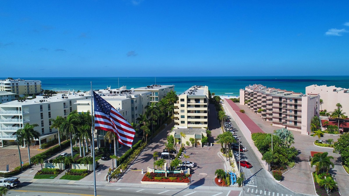 Aerial view reveals a beach community, highlighting well-maintained buildings amidst lush palm trees. The ocean is visible in the background, with sandy shores meeting calm waters. An American flag waves prominently in the foreground, symbolizing the area's coastal charm.