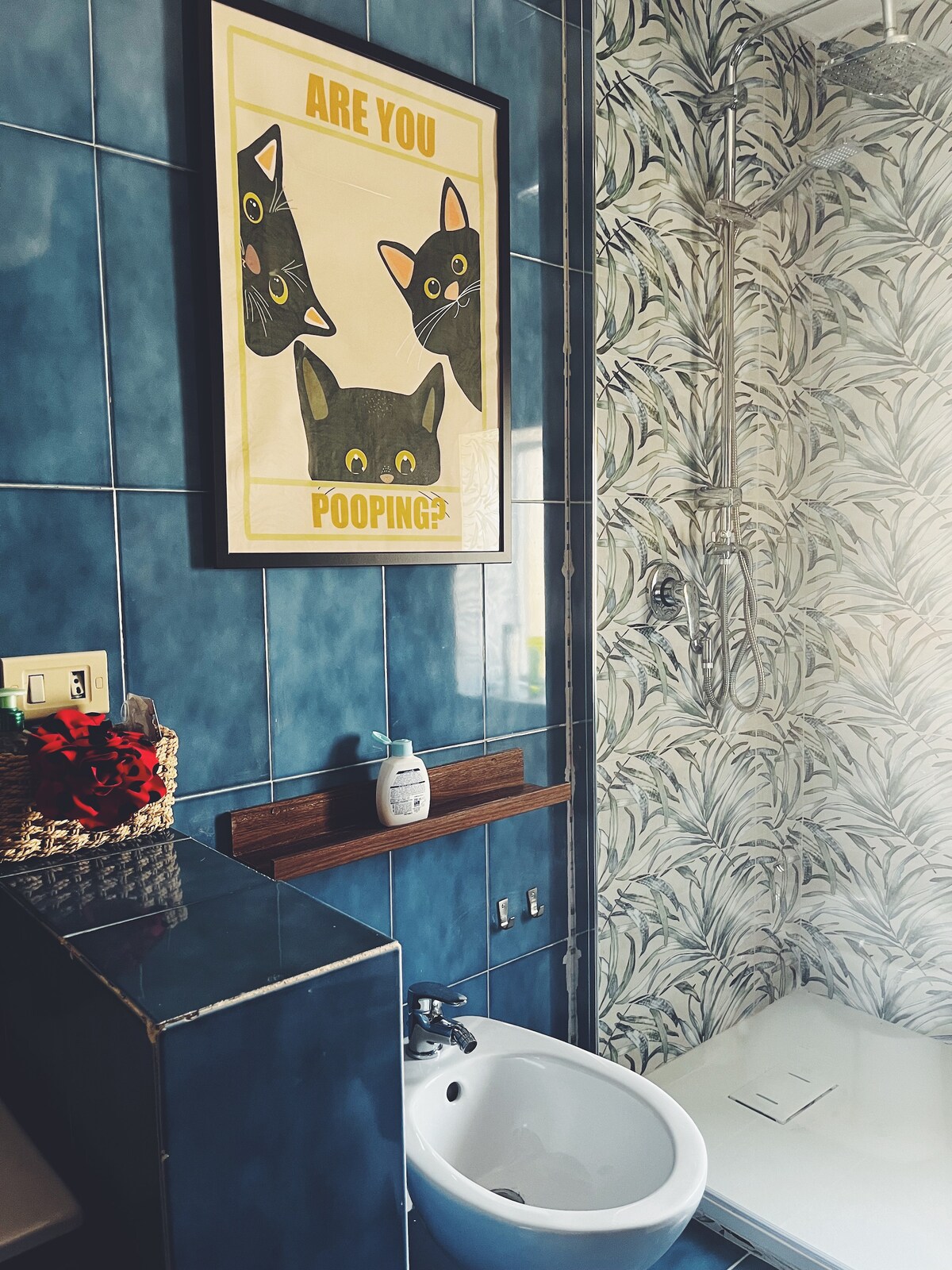 The bathroom features vibrant blue tiles complemented by a playful cat-themed wall art piece. A shower area with a glass door is visible, alongside a modern bidet and a minimalist sink. A patterned wallpaper adds a touch of character to the space.