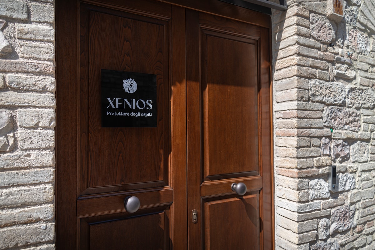 The entrance features a set of solid wooden doors with an elegant design, adorned with a prominent sign displaying 'Xenios' in a stylish font. The surrounding stone wall adds character, showcasing a blend of light and darker tones.