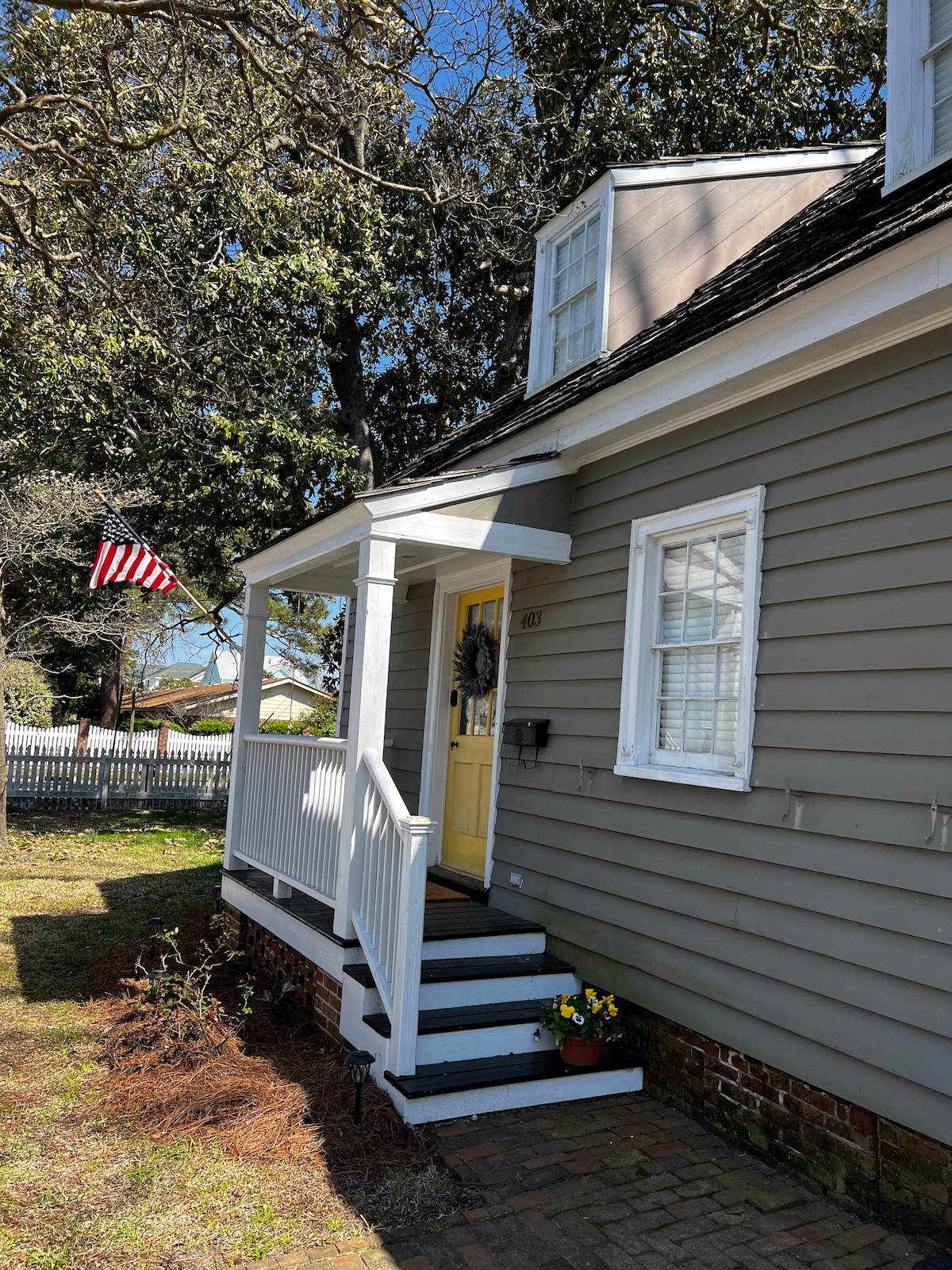 The exterior of a historic home is showcased, featuring light grey siding and a bright yellow door. A welcoming porch is accessed by white steps, complemented by a decorative wreath. An American flag is displayed nearby, surrounded by greenery and a flowering pot.