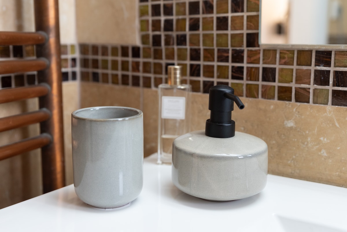 A clean bathroom countertop displays a grey ceramic cup and a pump dispenser, accompanied by a glass bottle. The backdrop features a mosaic tile design in various earthy tones, contributing to a modern and organized aesthetic.