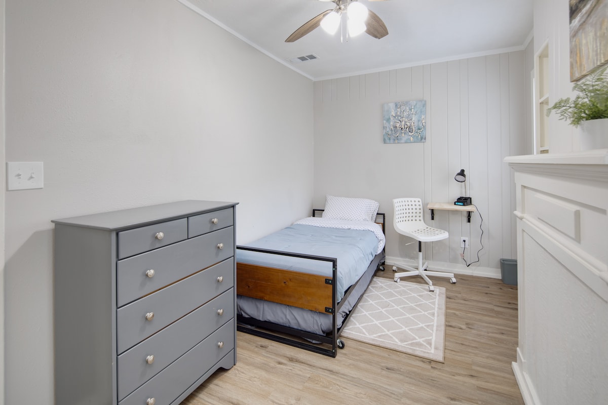 A cozy room features a twin bed with a simple, light blue comforter. A gray dresser stands against one wall, while a small desk with a chair is positioned nearby. Soft, warm lighting is provided by a ceiling fan, enhancing the inviting ambiance.