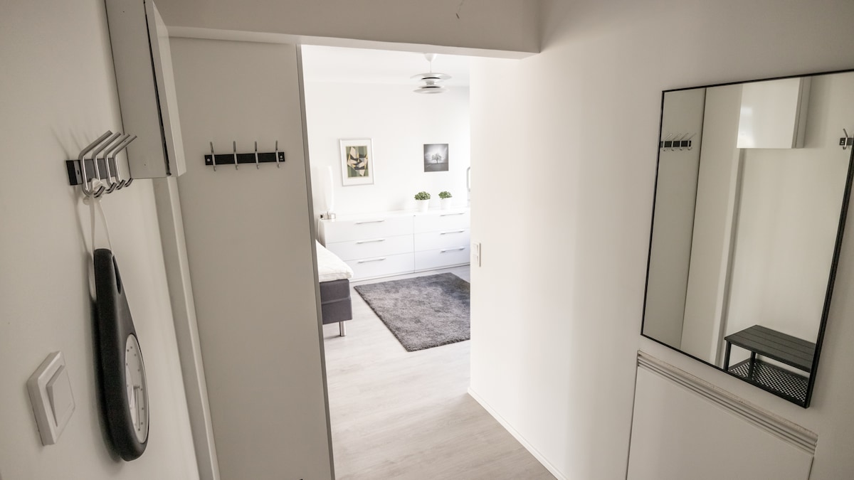 A hallway is shown leading into a well-lit living space. A large mirror is mounted on the wall, while minimalist decor is visible. A soft gray rug lies on the light-colored flooring, and a ceiling fan can be seen in the room beyond.