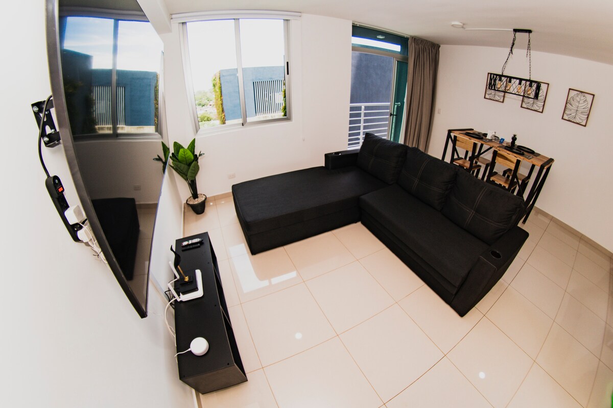 A modern living area is showcased with a sectional sofa placed adjacent to a mounted television. A dining table with six chairs is situated nearby. Large windows allow natural light to illuminate the space, while a small plant adds a touch of greenery.