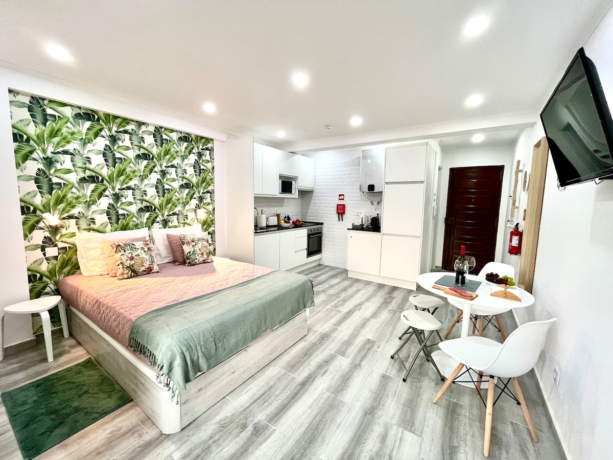A modern studio apartment is presented, featuring a spacious bed with soft bedding and pillows. The living area includes a small dining table with two chairs and a kitchenette. Bright lighting highlights the greenery wallpaper, while the floor is finished with light-colored planks.